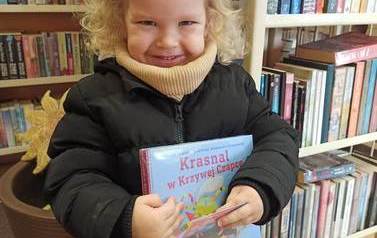 Uśmiechnięta dziewczynka o blond włosach i w czarnej kurtce pokazuje swoja nową książkę i kartę małego czytelnika.