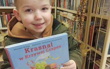 Chłopiec o blond włosach pokazuje swoją nową książkę. W tle regały z książkami.