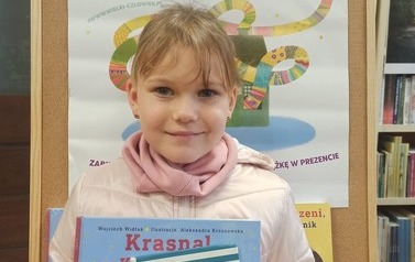 Dziewczynka o blond włosach pokazuje swoją nową książkę i książkę wypożyczoną. W tle plakat promujący akcję MAŁA KSIĄŻKA WIELKI CZŁOWIEK.