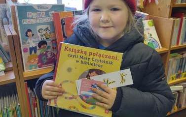 Dziewczynka w czerwonej czapeczce i czarnej kurtce pokazuje swoją nową książkę z kartą i zakładką do książki.