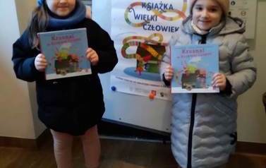 Dwie dziewczynki stoją przy tablicy z plakatem akcji "Mała Książka wielki człowiek" i pokazują swoje nowe książki.