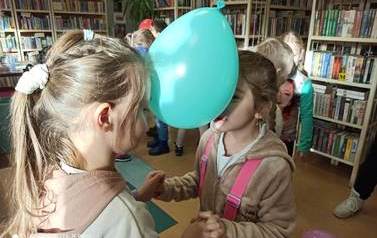Dwie dziewczynki stoją w parze twarzami do siebie i trzymając się za ręce trzymają głowami zielony balon.