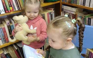 Jedna dziewczynka w blond włosach w warkoczyku siedzi na krzesełku i przegląda książeczkę, a druga stoi obok trzymając w ręku maskotkę pieska i zagląda do książeczki, kt&oacute;ra trzyma koleżanka.