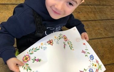 Chłopiec siedzi na podłodze, trzyma w dłoniach dużą kartke na kt&oacute;rej jest narysowany haft kociewski.