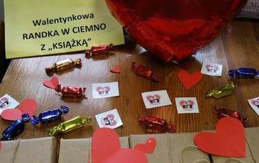 Na stoliku leżą książki zapakowane w brązowy papier owninięte sznurkiem i z przyklejonymi cytatami o miłości. Na nich rozsypane są serduszka, cukierki i karteczki z serduszkami. Obok jest duży balon w kształcie serduszka.