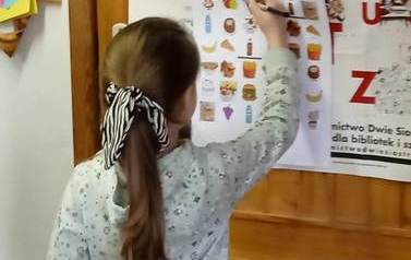 Dziewczynka zaznacza zdrowe produkty dla chorej babci na kartce przyczepionej do drzwi.