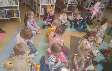 Dzieci siedzą na karimatach i bawią się pluszowymi książeczkami. Z tyłu Pani z Przedszkola trzyma na kolanach dziewczynkę.