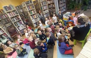 Widok z g&oacute;ry na siedzące dzieci na dywanie w bibliotece słuchające bajki czytanej przez bibliotekarkę.