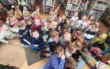 Widok z g&oacute;ry na siedzące dzieci na dywanie w bibliotece słuchające bajki.
