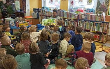 Dzieci siedzą na podłodze i patrzą na bibliotekarkę, kt&oacute;ra im pokazuje książeczkę o dinozaurach w formacie 3D.
