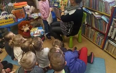 Dzieci siedzą na karimatach i patrzą na książeczkę przy kt&oacute;rej stoi dziewczynka, kt&oacute;rej bibliotekarka pokazuje coś z książeczki. Dziewczynka trzyma w rękach siateczkę i bawi się w robienie zakup&oacute;w z książeczki.