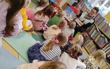 Dzieci siedzą na podłodze na karimatach i patrzą na bibliotekarkę, kt&oacute;ra pokazuje im kartę magnetyczną i książeczki kt&oacute;re wchodzą w skład wyprawki &quot;Małego Czytelnika&quot;