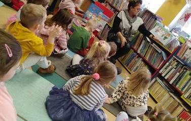 Dzieci siedzą na karimatach na podłodze i słuchają bajki o zimie czytanej przez bibliotekarkę.