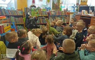 Dzieci siedzą na dywanie i słuchają bibliotekarki, kt&oacute;ra im czyta bajkę i pokazuje książeczkę o dinazaurach w formacie 3D. Jedna dziewczynka trzyma w rękach maskotkę dinozaura.