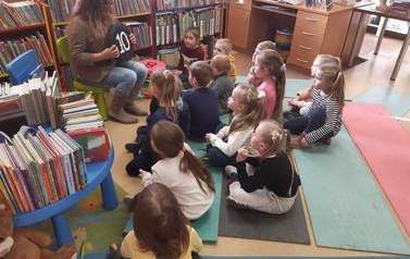 Dzieci siedzą na karimatach i patrzą na bibliotekarkę, kt&oacute;ra im pokazuje czarne, okrągłe siedzisko z numerem 10.