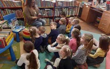 Dzieci siedzą na karimatach i słuchają bajki czytanej przez bibliotekarkę.