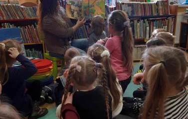 Dzieci siedzą na karimatach,a bibliotekarka czyta im bajkę w formacie 3D o dinozaurach pokazując przy tym ilustracje.