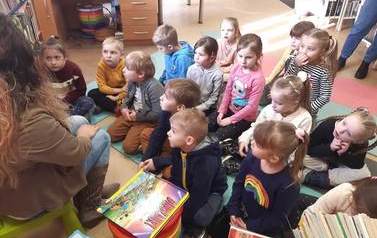 Dzieci siedzą na karimatach i patrzą na bibliotekarkę, kt&oacute;ra im coś opowiada. Z boku widać przygotowane książeczki do czytania.