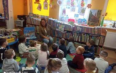 Dzieci siedzą na karimatach,a bibliotekarka czyta im bajkę pokazując przy tym ilustracje.
