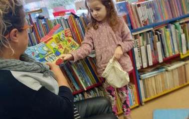 Bibliotekarka pokazuje książeczkę pluszową, a przy Niej stoi dziewczynka z siateczką aby zabawić się w robienie zakup&oacute;w z książeczki. Rączką odczepia z książeczki jakiś przedmiot.