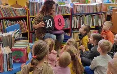Dzieci siedzą na karimatach,a bibliotekarka pokazuje im siedziska z numerami 10 i 8