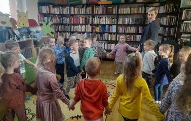 Dzieci wraz ze swoją Panią tańczą w dużym k&oacute;łku śpiewając. W środku koła tanczą dwaj chłopcy.