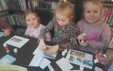 Trzy dziewczynki o blond włosach naklejają bajkowe puzzle na białą kartkę. Jedna z dziewczynek trzyma w ręku klej i patrząc w aparat się uśmiecha.