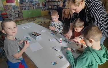 Czterech chłopc&oacute;w stoi przy białym stoliku i nakleja na białą kartkę puzzle. Jednemu z chłopc&oacute;w pomaga Pani Przedszkolanka.
