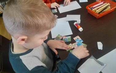 Chłopiec o blond włosach układa przy stole papierowe puzzle z bajki "trzy małe świnki"