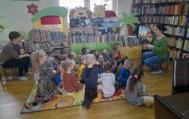 Na dywanie siedzą dzieci i słuchają bajki czytanej przez bibliotekarkę. Z lewej strony z tyłu siedzi na krzesełku Pani opiekunka.