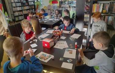 Przy dw&oacute;ch dużych stołach siedzą dzieci i naklejają na białą kartkę puzzle z bajki "Trzy małe świnki".