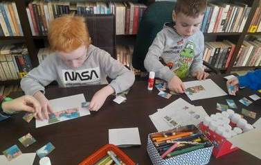 Dw&oacute;ch chłopc&oacute;w przy stoliku nakleja na białą kartkę puzzle z bajki "Trzy małe świnki".