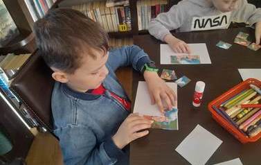Dw&oacute;ch chłopc&oacute;w przy stoliku nakleja na białą kartkę puzzle z bajki "Trzy małe świnki".