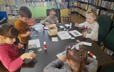 Dzieci siedzą przy stoliku i naklejają na kartkę puzzle z bajki &quot;Trzy małe świnki&quot;.