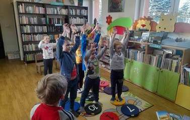 Dzieci stoją na dywanie i wyciągają się z uniesionymi rękoma w g&oacute;rę naśladując bibliotekarkę.