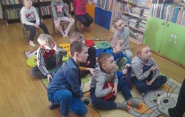 Dzieci siedzą na krzesełkach i siedziskach i słuchają bajki, ktorą czyta im bibliotekarka.