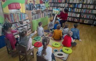 Dzieci siedzą na krzesełkach i siedziskach i słuchają bajki, ktorą czyta im bibliotekarka. Jeden chłopiec wskazuje coś rączką w na ilustrację, kt&oacute;rą pokazuje bibliotekarka.