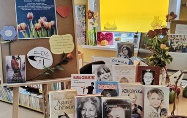 Na biurku są wystawione książki o kobietach, cukierki i aforyzmy. Z lewj strony stoi tablica przystrojona  w plakat z życzeniami dla kobiet, cytatami i r&oacute;żą.