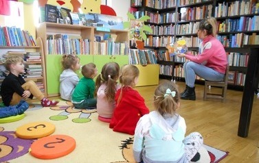 Dzieci siedzą na siedziskach i słuchają bajki czytanej przez bibliotekarkę, kt&oacute;re siedzi przed nimi na krzesełku.