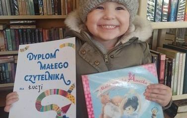 Bardzo uśmiechnięta dziewczynka pokazuje sw&oacute;j plakat i wypożyczoną książkę.
