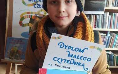 Chłopiec pokazuje sw&oacute;j dyplom i wypożyczoną książkę. W tle widac plakat z akcji Mała Książka Wielki Człowiek
