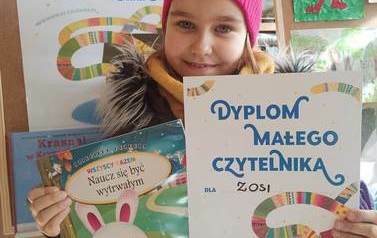 Dziewczynka pokazuje dyplom i wypożyczoną książkę. W tle widac plakat akcji Mała Książka Wielki Człowiek