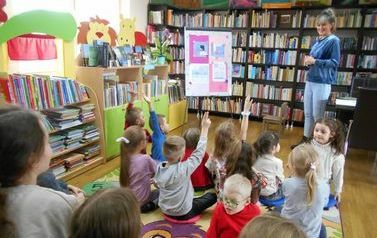 Dzieci siedzą na dywanie i zgłaszają się wyciągając ręce do g&oacute;ry. Bibliotekarka stoi przed nimi i uśmiecha.