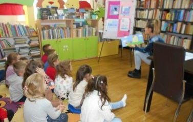 Dzieci siedzą na dywanie i słuchają bajki czytanej przez bibliotekarkę. Z boku stoi tablica metalowa na kt&oacute;rej przyczepione są zadania to tekstu z bajki.
