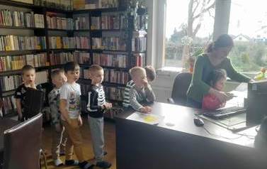 Dzieci stoją w kolejce przy biurku aby skorzystać z czytnika,a bibliotekarka pomaga dziecku przyłożyć książkę do czytanika.