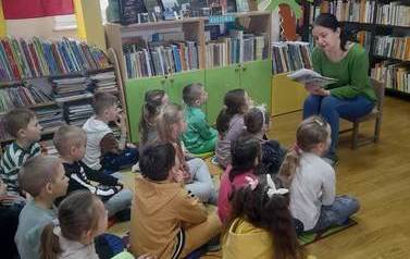 Dzieci siedzą na dywanie i patrzą w stronę bibliotekarki, kt&oacute;ra czyta im bajkę.