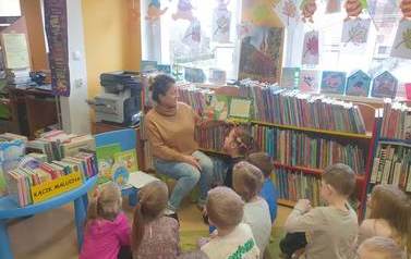 Na dywanie siedzą dzieci i słuchają bajki czytanej przez bibliotekarkę. 