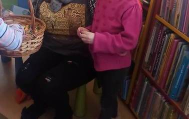 Mała dziewczynka o blond włosach stoi przy bibliotekarce, kt&oacute;ra trzyma koszyk z cukierkami. W koszyku widać czyjąś rączkę.
