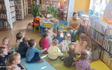 Dzieci siedzą na dywanie i karimatach skierowane do bibliotekarki czytającej im bajkę.