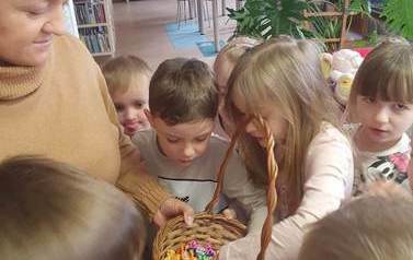 Bibliotekarka stoi z koszykiem cukierk&oacute;w otoczona częstującymi się dziećmi.